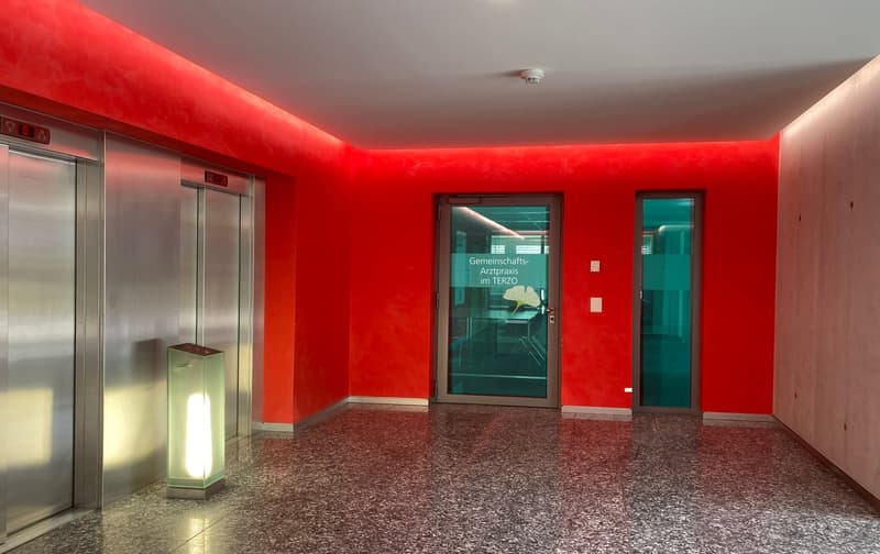 The image shows a brightly lit interior space with red walls and a terrazzo floor. There are two glass doors leading to another room, and an elevator can be seen on the left side of the image. The space appears to be a medical or professional office, as i