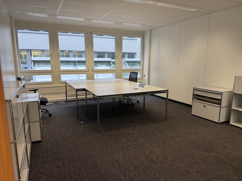 An empty office with a large table, two chairs, and a shelf with drawers. The room has white walls, a white ceiling, and a carpeted floor. There are large windows with blinds and a view of the outside.