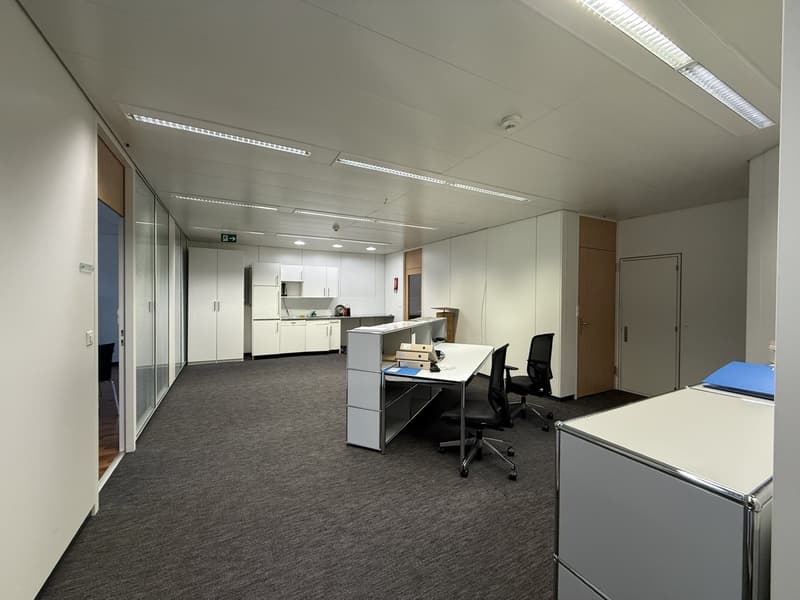 Empty office space with white walls, gray carpet, white desks, black office chairs, white cabinets, and ceiling lights.