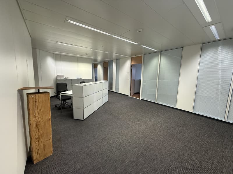 Empty office with white walls, gray carpet, white desk, black office chair, white cabinets, glass doors, ceiling lights