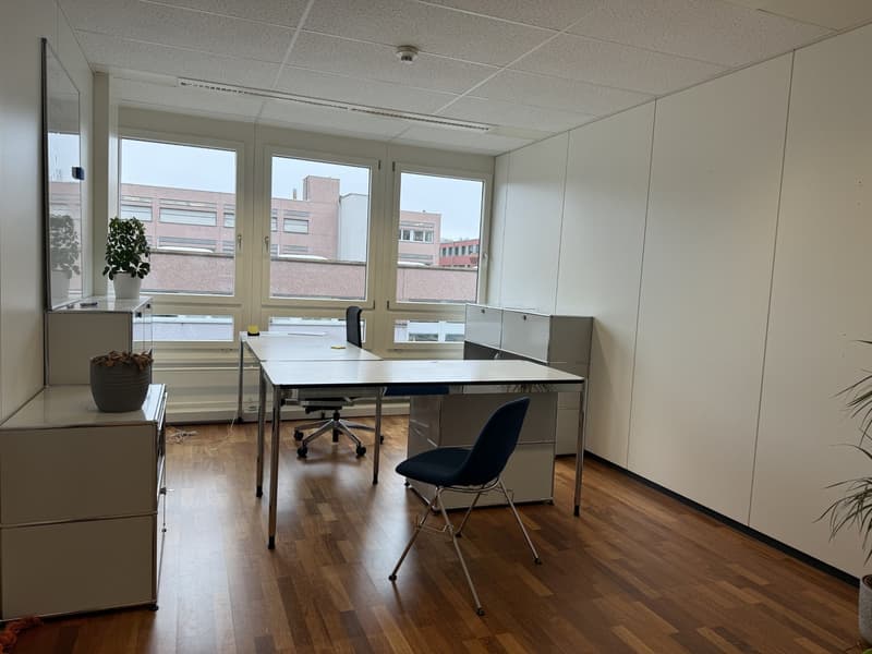 An empty office room with a desk, two chairs, and a cabinet. The room has a wooden floor and white walls. There are two large windows with a view of the outside. There are two potted plants on the left side of the room.