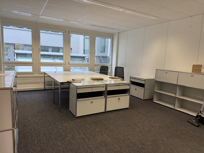 empty office with large windows, white walls, carpeted floor, desks, chairs, cabinets, and a vacuum cleaner