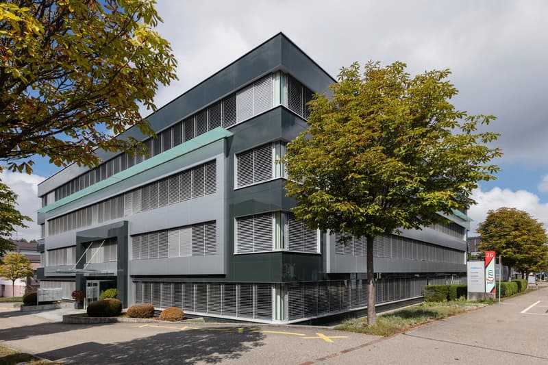 modern office building, gray exterior, multiple windows, entrance with canopy, parking area, trees, shrubs, signboard