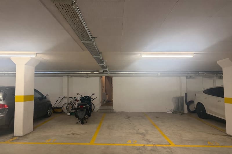 Underground parking with two cars and a motorcycle parked, yellow lines marking parking spots, white walls, and ceiling lights