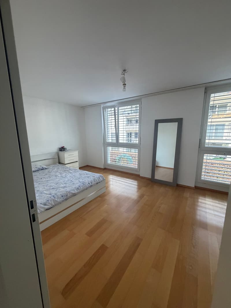 A bedroom with a large bed, a white cabinet, a mirror, wooden floors, and a window with a view of another building.