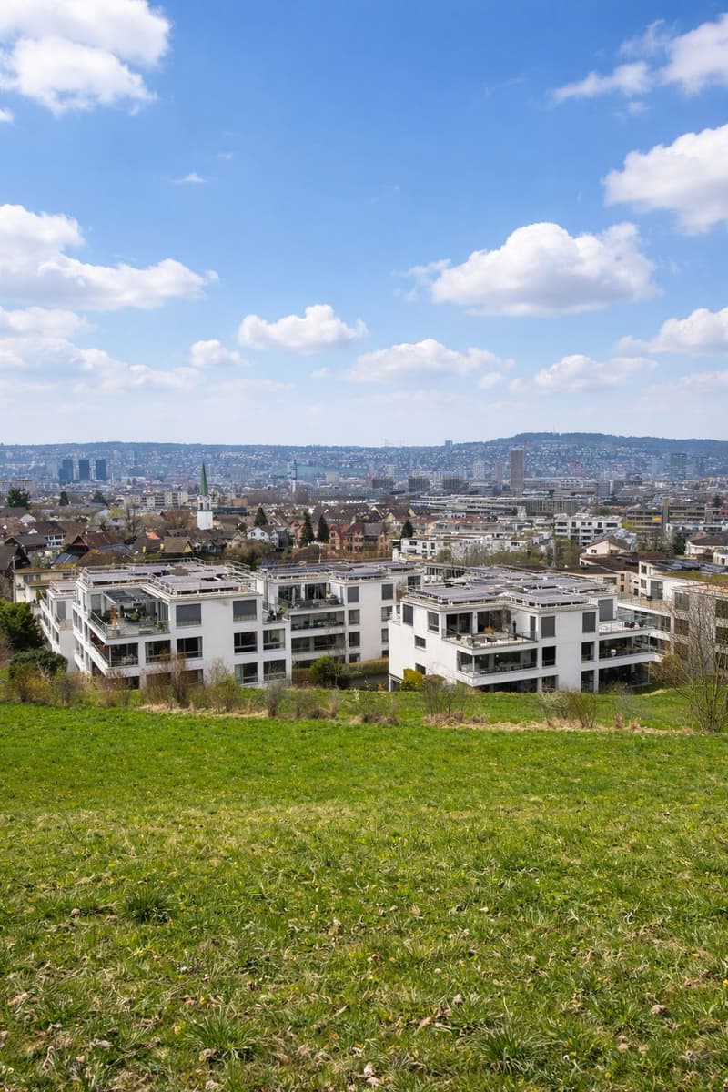 Aerial view of a modern apartment building with solar panels on the roof, surrounded by a green field and a cityscape in the background.