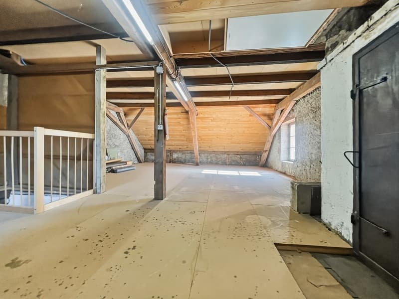 A room under the roof with wooden beams, unfinished floor, an entrance with a staircase, and a wall with a window.