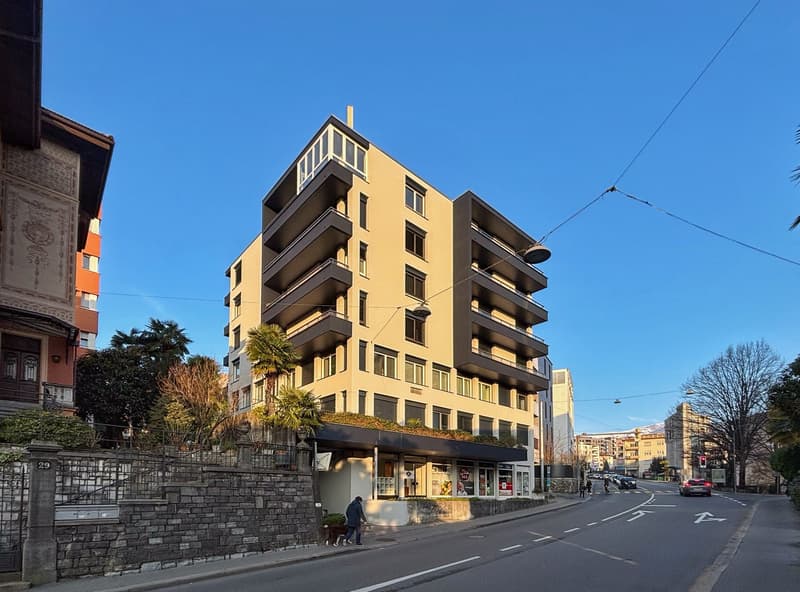 Modern multi-story building with balconies, located on a street with a sidewalk, greenery, and a road with cars and pedestrians.