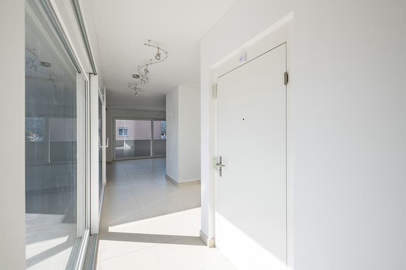 Empty hallway with white tiles, glass doors, and a balcony visible through the doors.