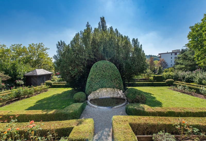 well-manicured garden with a central fountain, neatly trimmed hedges, and a variety of plants and flowers