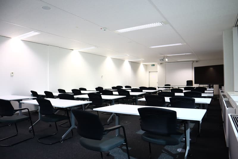 Classroom with white walls, white desks, black chairs, projector screen, and ceiling lights.