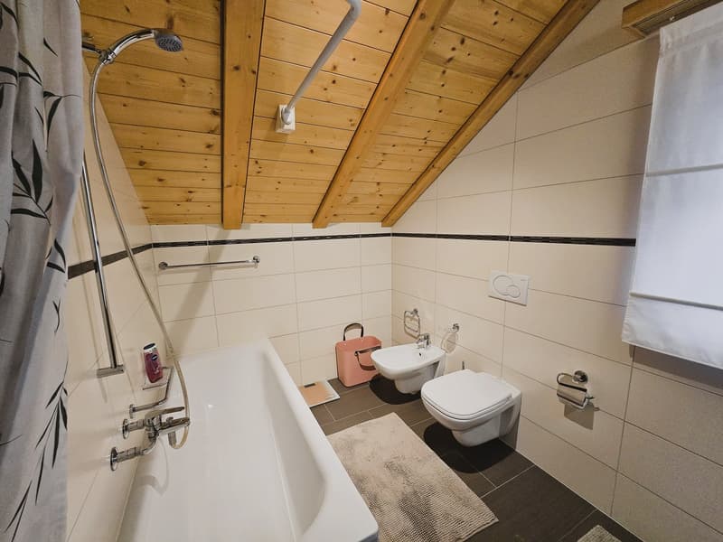 Bathroom with wooden ceiling, white bathtub, white toilet, bidet, white tiles, shower head, shower curtain, rug, and a pink basket.