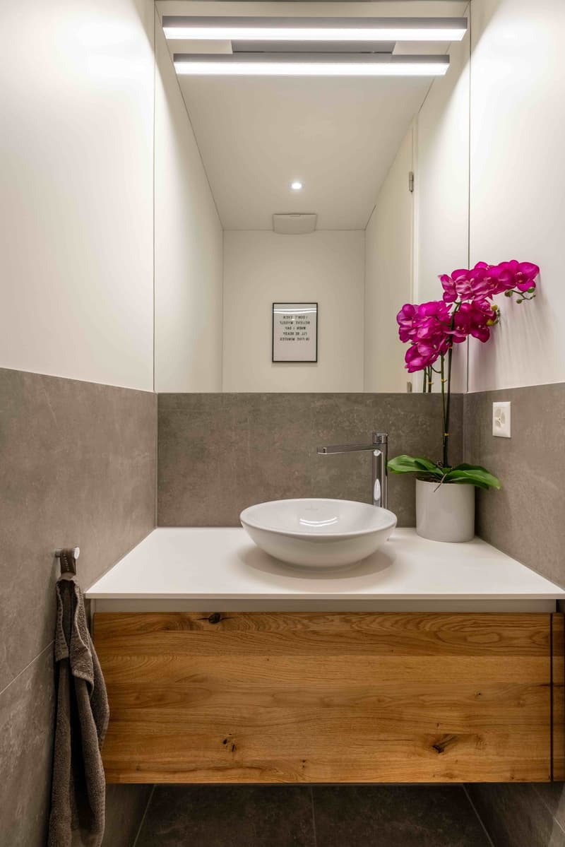 Modern bathroom, grey tiles, wooden vanity, white sink, modern faucet, flower in a white vase, mirror, towel holder, recessed lighting