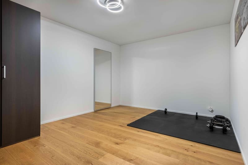An empty room with white walls, hardwood floors, a wardrobe, a mirror, a workout mat, and exercise equipment