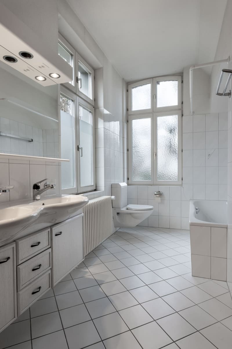 White tiled bathroom, white cabinets, white sink, white toilet, bathtub, double window