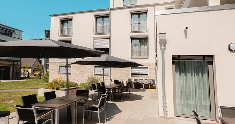 Outdoor dining area with tables and chairs, large umbrellas, brick wall, windows, entrance door
