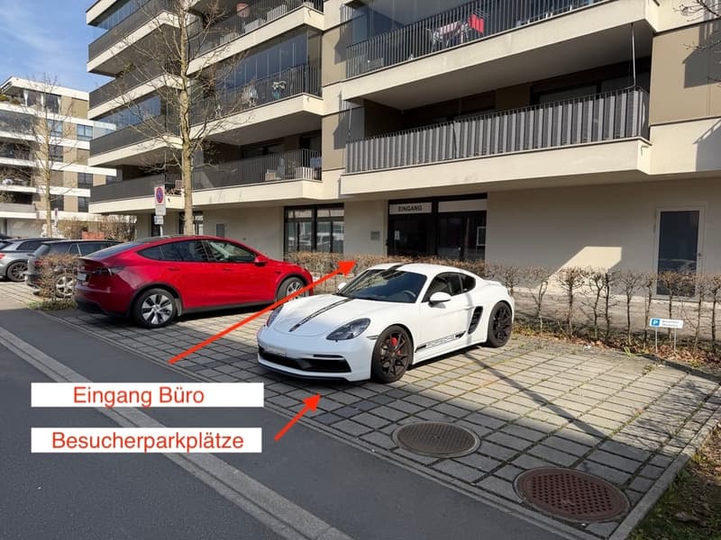 A multi-story building with glass windows and balconies. Two cars are parked in front of the building, one is a red Tesla and the other is a white Porsche. The building has a sign that reads 'Eingang' and 'Besucherparkplatze'. There are bushes and a tree
