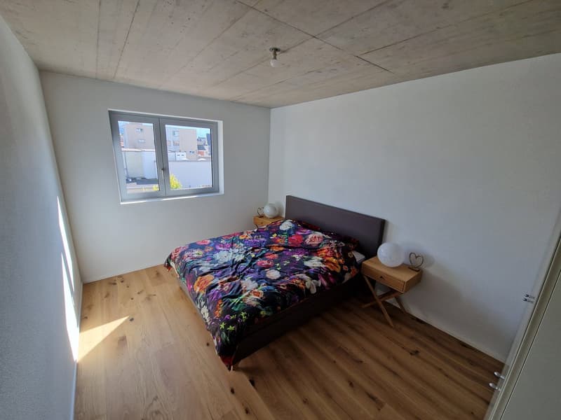 Bedroom with wooden floor, white walls, a bed with a floral cover, two nightstands with lamps, and a window.