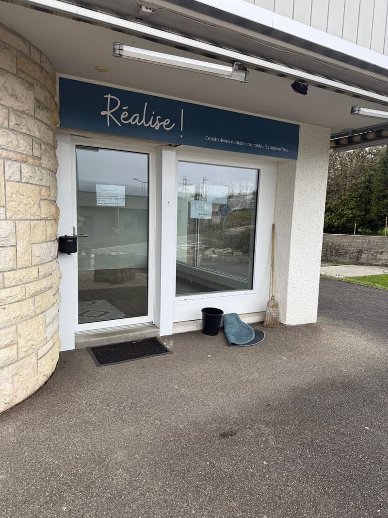 Entrance to a building with a sign that reads 'Realise!', glass doors, a black mat, a broom, and a black bucket.