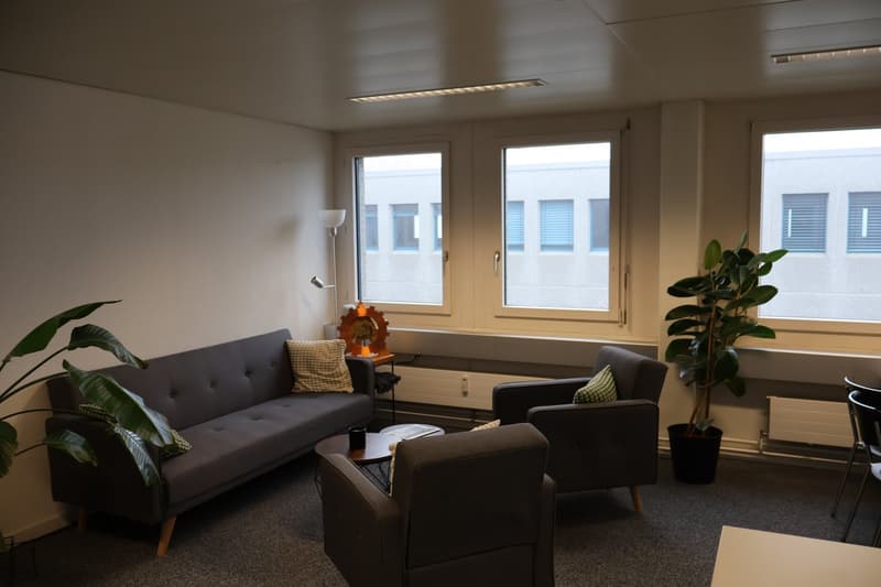 A room with a couch, two armchairs, a coffee table, a floor lamp, and a plant. There are three windows with views of the outside.