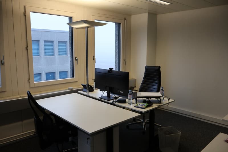 Two desks, two chairs, computer monitor, lamp, trash bin, water bottles, building view