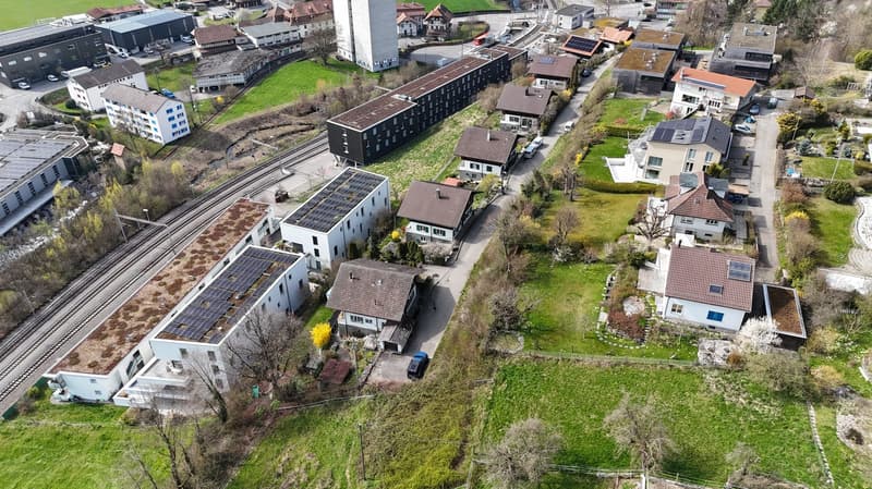 Aerial view of a residential area with several houses and buildings, some with solar panels on the roofs, surrounded by green grass and trees, with a road and a railway track in the area.