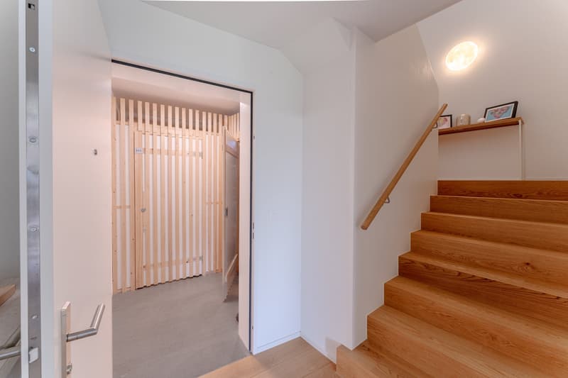 Entrance hall with wooden stairs, wooden flooring, white walls, wooden door, wooden railing, wooden partition, and a moon-shaped light fixture on the wall.