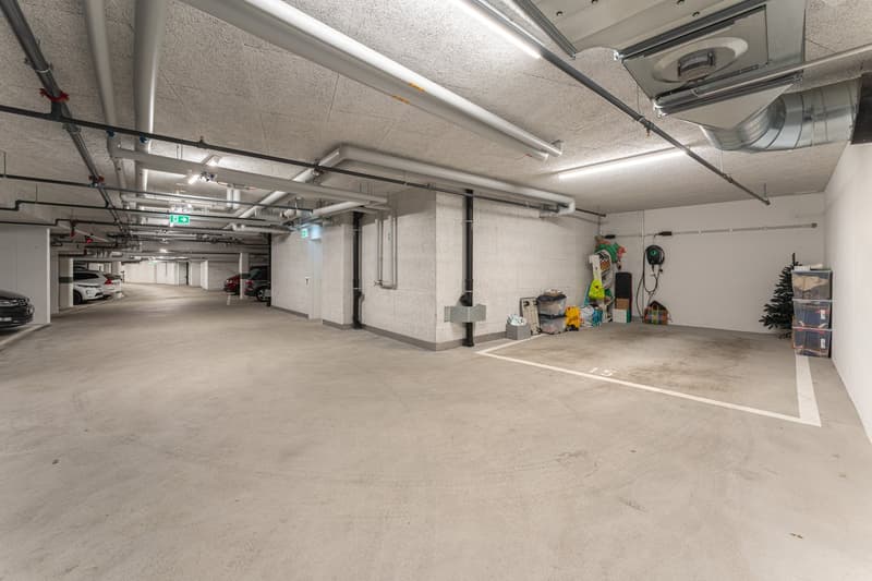 underground parking lot, concrete floor, white walls, ceiling with pipes and lights, empty parking spaces, storage boxes, electric vehicle charging station