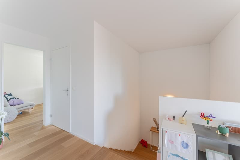 Empty room with white walls, wooden floor, white door, small desk, and a staircase leading to another room