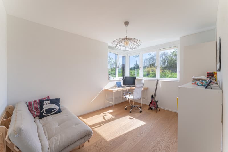 A room with a desk, chair, guitar, couch, and windows with a view of the outside.