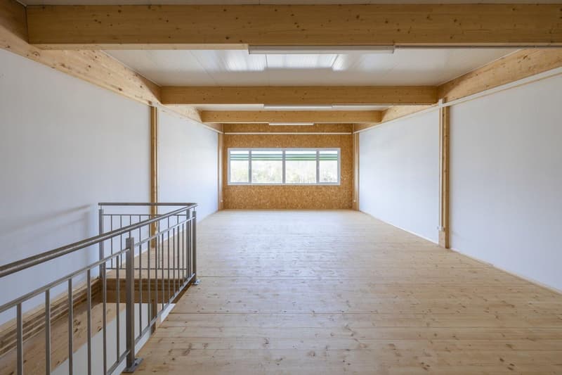 Large open space with wooden beams and ceiling, white walls, and hardwood flooring. There is a metal railing on the upper level overlooking the room below.