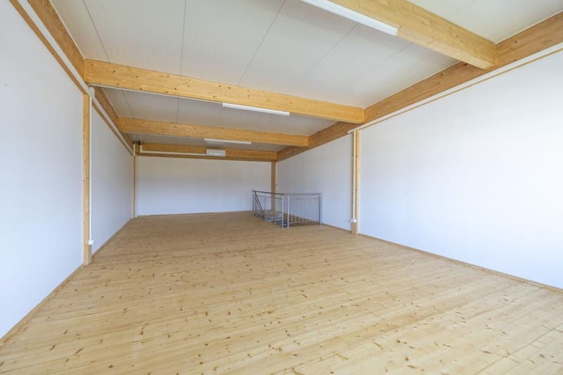 Large open space with wooden ceiling beams and a wooden floor. The room appears to be empty, with a metal railing visible in the background.