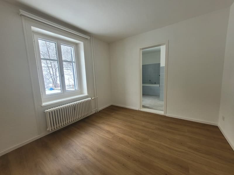 Empty room with white walls, wooden floor, large window, radiator.