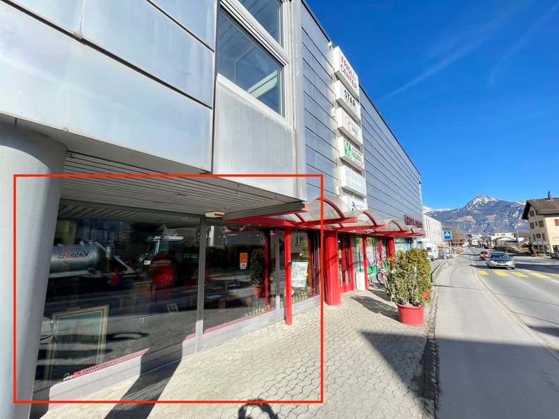 Commercial building with glass windows, red pillars, and a red awning. Potted plants and a bicycle are on the sidewalk. A car is driving on the road.