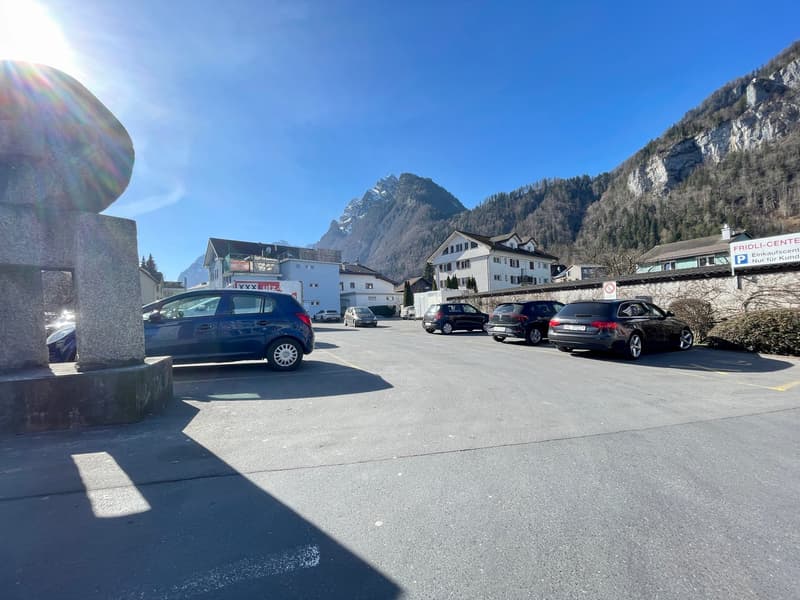 Parking lot with several cars parked, surrounded by buildings and mountains