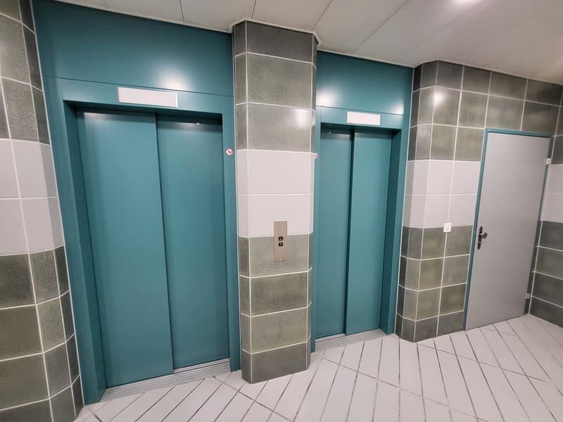 The image shows an elevator lobby with two elevator doors in a teal and gray color scheme. The walls are tiled with gray and teal tiles, and the floor is covered with white and gray tiles in a geometric pattern. The elevator doors have a modern, minimalis