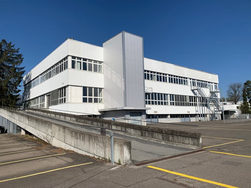 A large, multi-story industrial or commercial building with a white exterior and numerous windows. The building has a rectangular, modern design with a flat roof. There is a concrete platform or ramp leading up to the building, and yellow lines on the gro