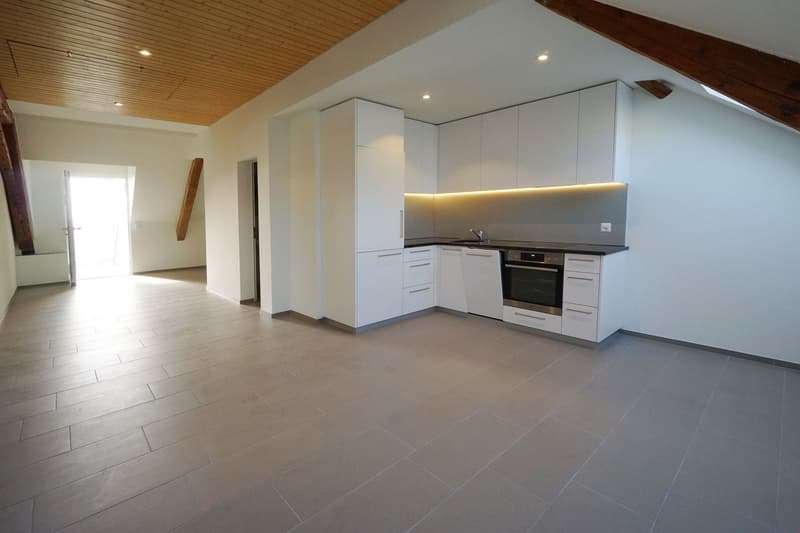Modern, open-concept kitchen with white cabinets, built-in appliances including a dishwasher, and gray tile flooring. The kitchen is integrated into a larger living space with a vaulted ceiling and exposed wooden beams.