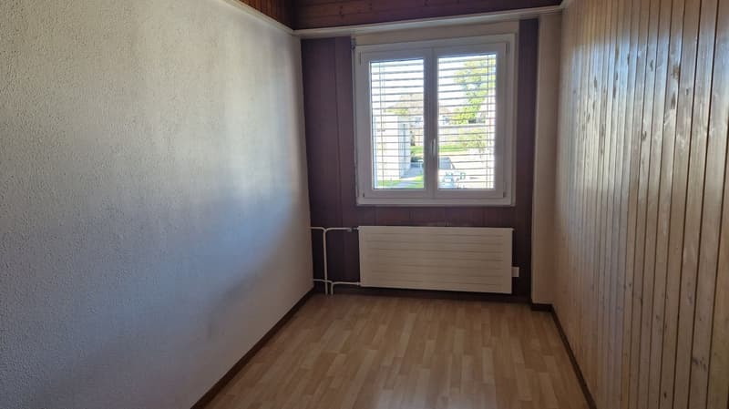 Empty room with wooden walls, a window with white shutters, a white radiator, and hardwood floors.