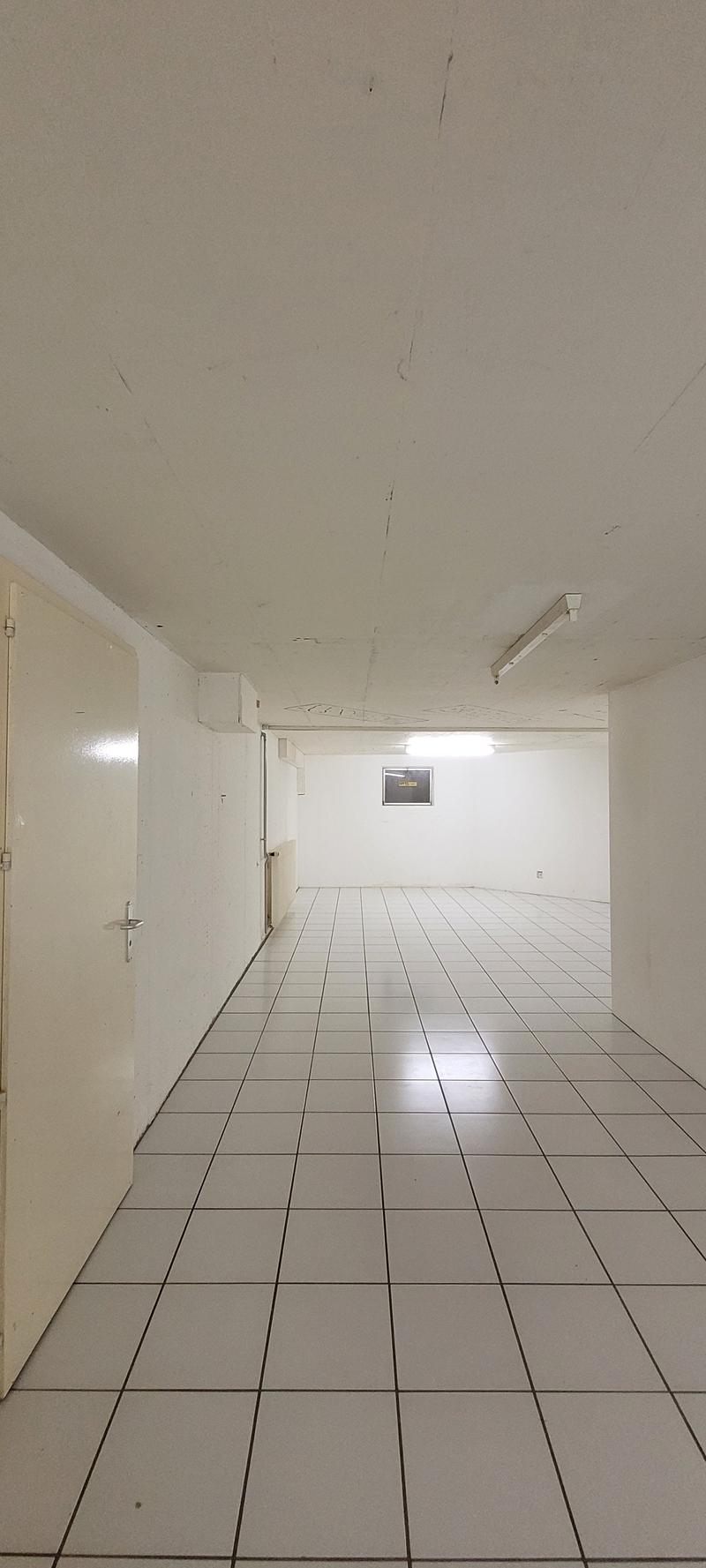 The image shows a long, empty hallway with white walls and a tiled floor. The hallway appears to be in a commercial or institutional building, with a door visible at the end of the hallway.