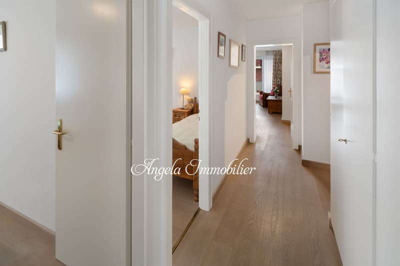 Empty hallway with wooden floor, white walls, wooden bed, nightstand, lamp, framed pictures on the wall, open door leading to another room with a couch and a table.