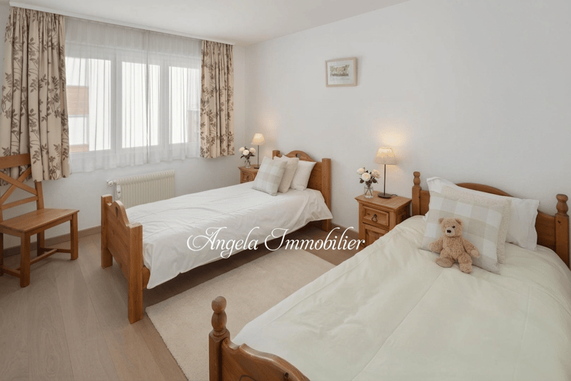 Two single beds, wooden chair, wooden nightstands, lamps, white curtains, white walls, wooden floor, teddy bear