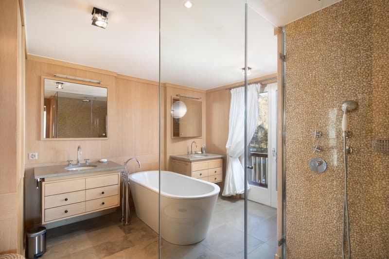modern bathroom with wooden walls, white bathtub, double sink, glass shower enclosure, mirror, wooden cabinets, and a trash bin