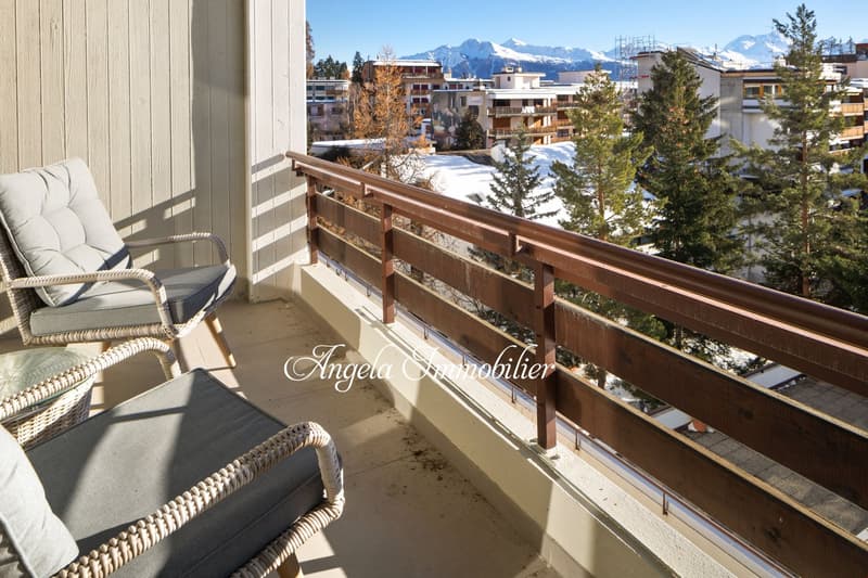 balcony with wooden railing, beige wicker chairs, wooden floor, mountainous view, snowy ground, trees, buildings