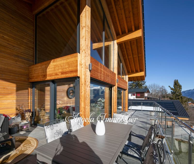 Wooden house with large windows, balcony, and outdoor seating area with a table and chairs