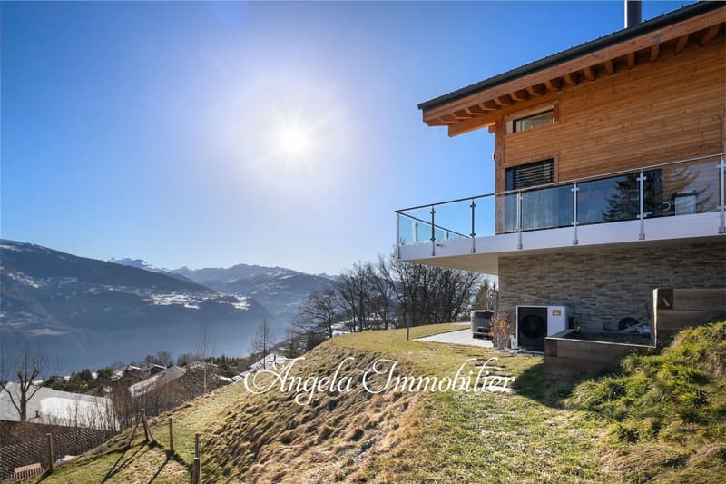 Wooden chalet-style house with a balcony overlooking a scenic mountain landscape. The house has large windows and a glass railing on the balcony, providing a panoramic view of the surrounding mountains and valley below.