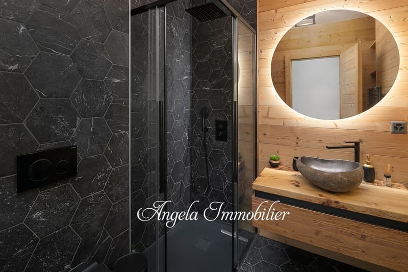 Luxurious bathroom with black hexagonal tile walls, a round backlit mirror, a wooden vanity with a stone vessel sink, and a glass shower enclosure.