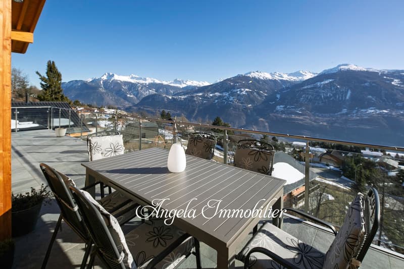 Outdoor terrace with a wooden table and chairs, overlooking a scenic mountain landscape with snow-capped peaks in the distance. The terrace has a glass railing and appears to be part of a larger property or building.