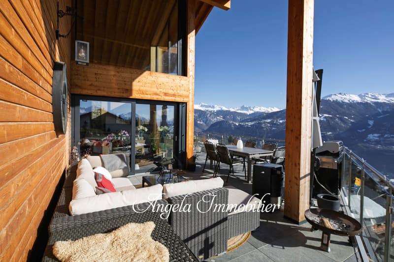 Wooden chalet with large windows and balcony overlooking snowy mountains, outdoor seating area with wicker furniture, cozy interior with sofa and decor