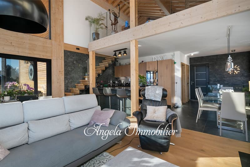 Spacious open-concept living area with wooden beams, stone wall, grey sofa, armchair, dining table and chairs, kitchen with bar stools, and a balcony or terrace visible through the large windows
